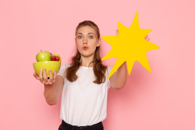 Бесплатное фото Вид спереди молодой женщины в белой футболке, держащей тарелку с фруктами и большим желтым знаком на розовой стене