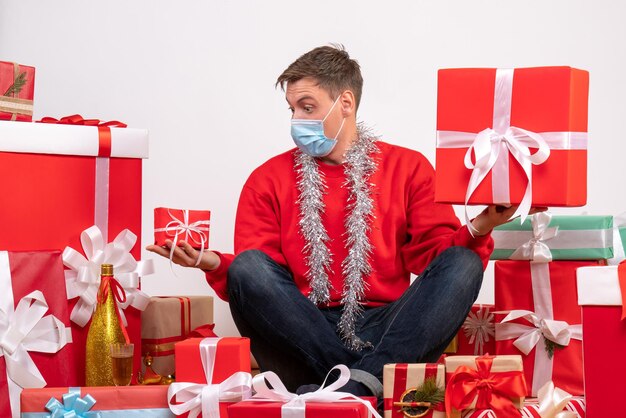 無料写真 白い壁にクリスマスプレゼントの周りに座っている滅菌マスクの若い男の正面図