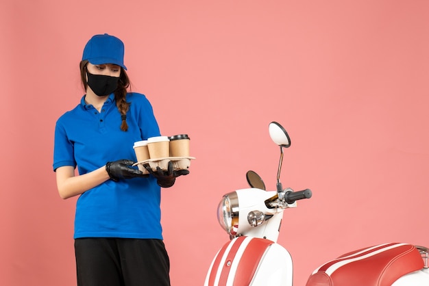 無料写真 パステル ピーチ色の背景にコーヒーの小さなケーキを保持しているオートバイの隣に立っている医療マスク手袋を着た若い驚いた宅配便の女の子の正面図