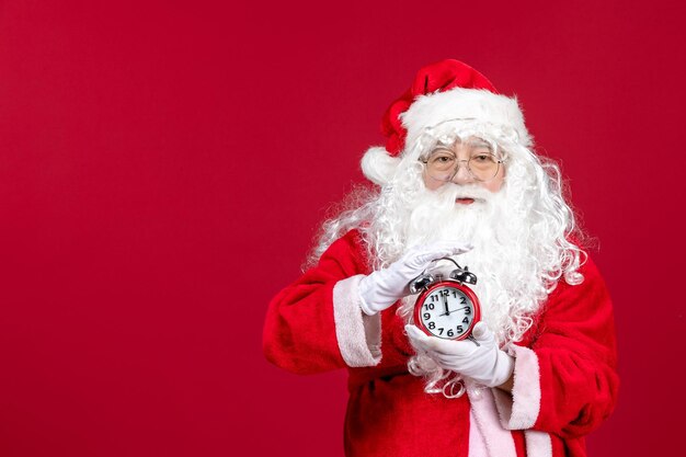 Front view of old santa claus in red suit holding clock on red wall