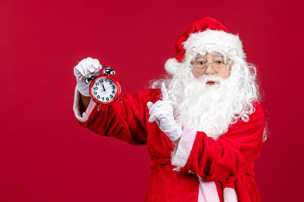 Front view of old santa claus in red suit holding clock on a red wall