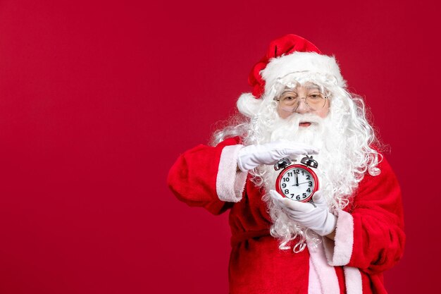 Front view of old santa claus in red suit holding clock on red wall