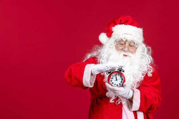 Front view of old santa claus in red suit holding clock on red wall