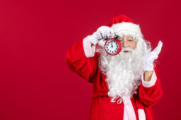 Front view of old santa claus in red suit holding clock on red wall