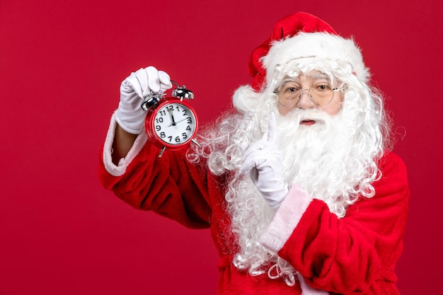 Front view of old santa claus in red suit holding clock on the red wall