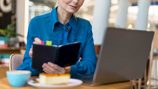 Front view of older business woman with glasses writing in agenda and looking at laptop