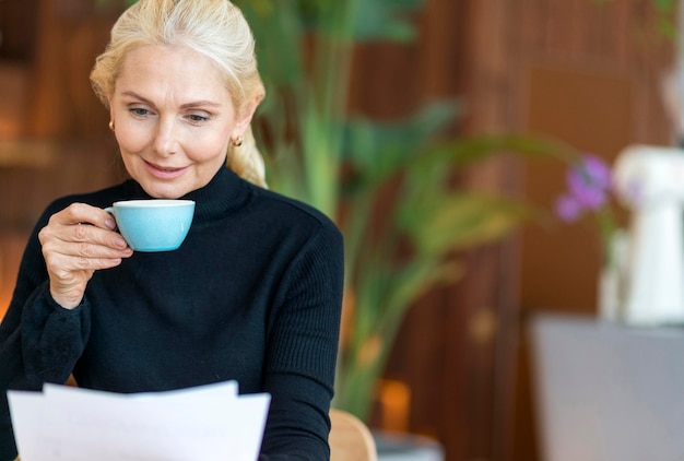 Front view of older woman at work reading papers while having coffee