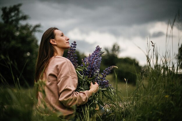 Front view of one pretty girl holding enormous bouquet of wild violet lupines, dressed in casual clothes on cloudy day