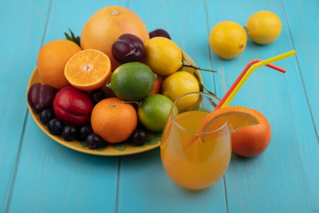 ターコイズブルーの背景に黄色いプレートにライムとチェリープラムオレンジプラムレモンとグラスの正面図オレンジジュース