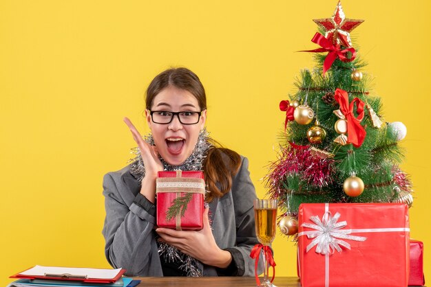 그녀의 행복 크리스마스 트리와 선물 칵테일을 보여주는 테이블에 앉아 안경으로 전면보기 기뻐 소녀