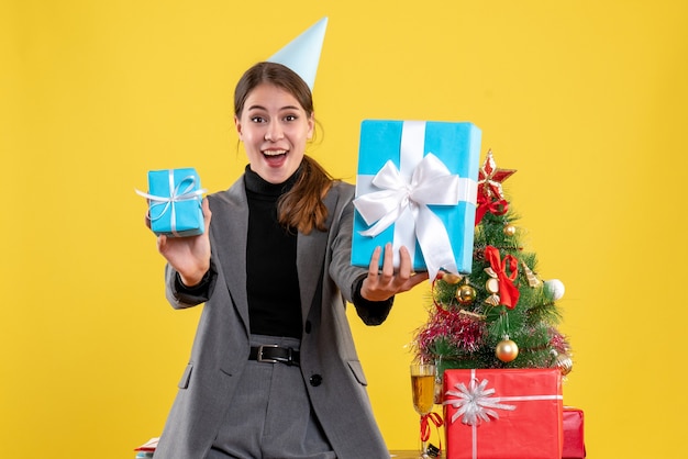 Front view overjoyed girl with party cap holding xmas gifts near xmas tree and gifts cocktail