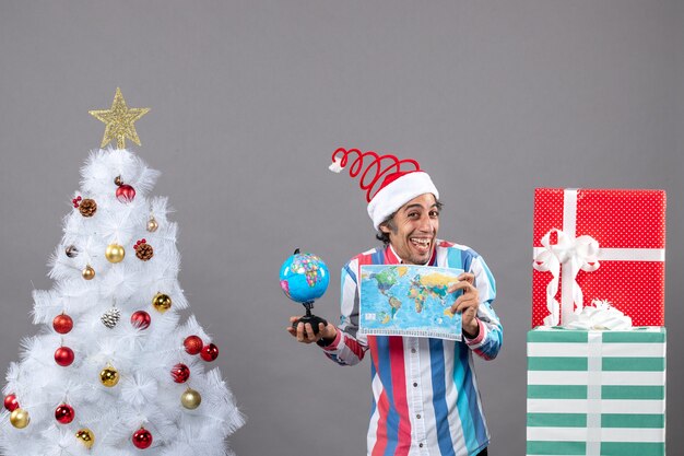 Front view overjoyed man with spiral spring santa hat holding world map and globe around xmas tree and presents