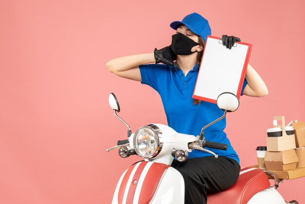 医療用マスクと手袋をはめ、パステル調の桃の背景に空の紙シートを持ったスクーターに座って注文を配達する、疲れきった宅配便の女性の正面図