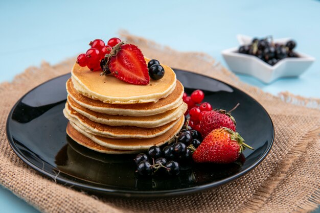 Front view of pancakes with red and black currants and strawberries on a black plate on a beige napkin