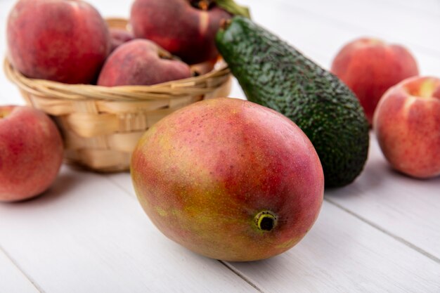 Front view of peaches on a bucket with mango avocado on a white surface