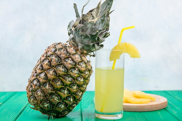 Vista frontale del succo di ananas con fette di ananas sul tagliere e ananas su superficie verde e superficie bianca