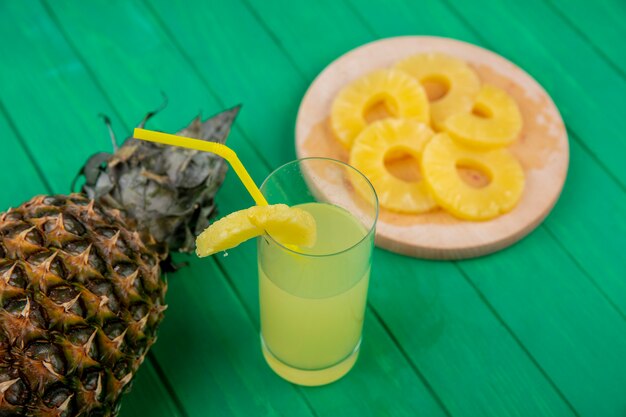 Front view of pineapple juice with pineapple slices on cutting board and pineapple on green surface