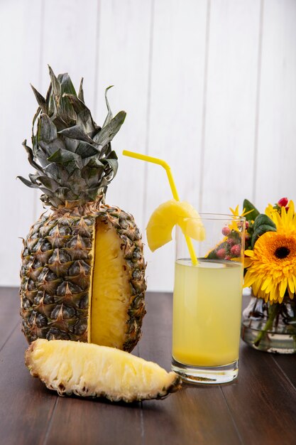 Front view of pineapple and pineapple slice and juice in glass with drinking tube and flowers on wooden surface