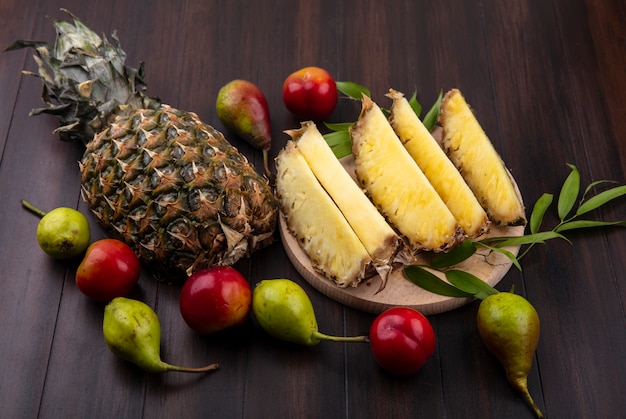 Front view of pineapple slices in plate with whole one and peach plum on wooden surface
