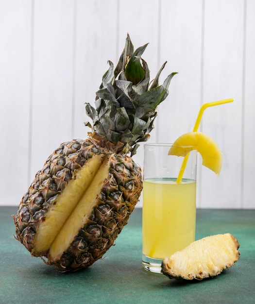 Front view of pineapple with one piece cut out from whole fruit and pineapple juice on green surface and white surface
