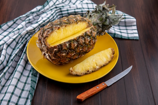 Front view of pineapple with one piece cut out from whole fruit in plate on plaid cloth with knife on wooden surface