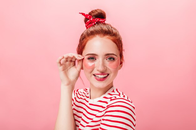 Front view of playful ginger girl with eye patches. Studio shot of young woman doing skincare treatment with smile.