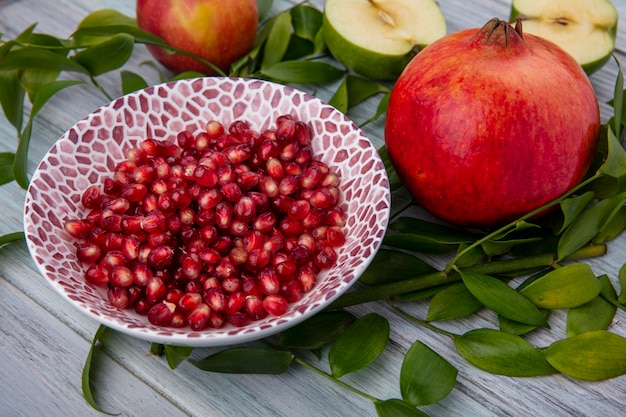 Front view of pomegranate berries in bowl and half cut apple with whole ones and leaves on wooden surface