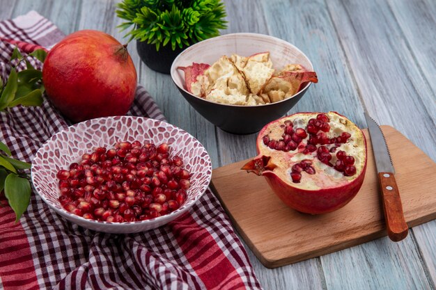 Front view of pomegranate berries in bowl with whole one and leaves on plaid cloth and half one with knife on cutting board with bowl of pomegranate shell on wooden surface