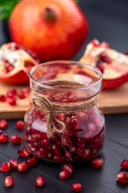 Front view of pomegranate berries in glass jar with whole and cut one on cutting board on black surface