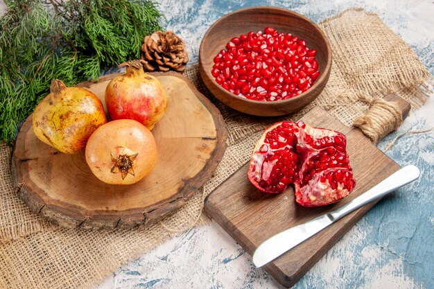 Front view pomegranates on round cutting board pomegranate seeds in bowl a cut pomegranate on chopping board pine tree branch on blue-white background
