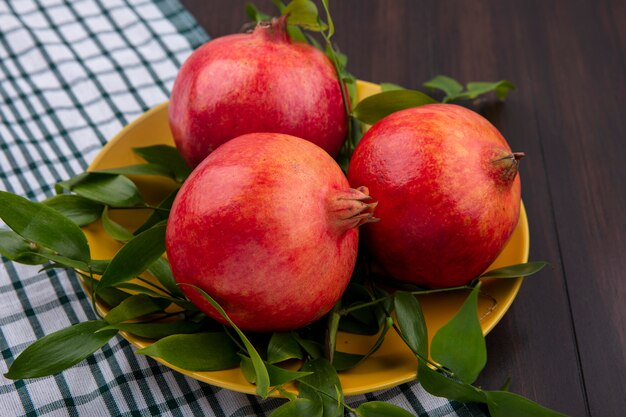 Front view of pomegranates with leaf branches on a yellow plate on a wooden surface