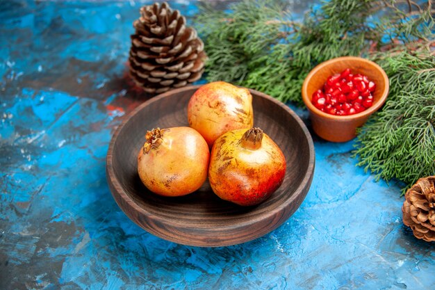 Front view pomegranates on wooden plate pomegranate seeds in wooden bowl pine tree branch and cones on blue