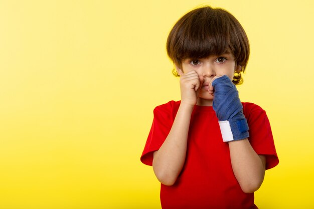 A front view posing cute boy in red t-shirt and blue tissue glove on the yellow wall