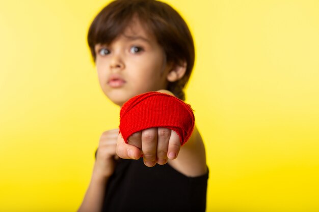 A front view posing little kid in black t-shirt and with one hand tied with red tissue on the yellow wall