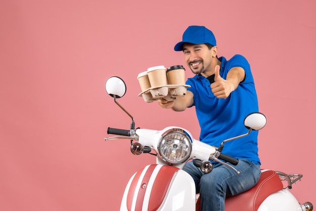 Front view of positive courier man wearing hat sitting on scooter making ok gesture on pastel peach background