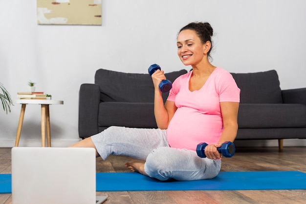 Front view of pregnant woman at home exercising on mat with laptop and weights