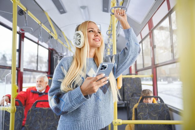 Front view of pretty blonde girl with phone looking at window smiling standing inside moving bus