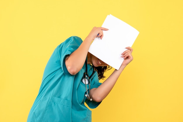 Front view of pretty female doctor covering her face with papers on yellow wall