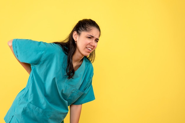 Front view of pretty female doctor holding her back with pain on yellow wall