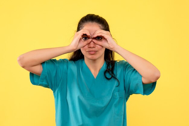 Front view of pretty female doctor making hands binoculars on yellow wall