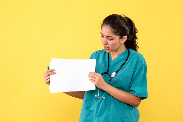 黄色の壁に立っている紙を保持している制服を着たきれいな女性医師の正面図
