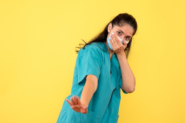 Front view of pretty female doctor with medical mask covering her mouth on yellow wall