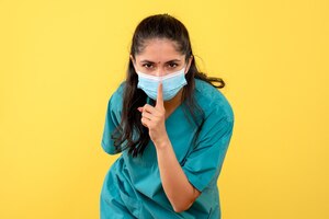 Front view of pretty female doctor with medical mask making shh sign on yellow wall