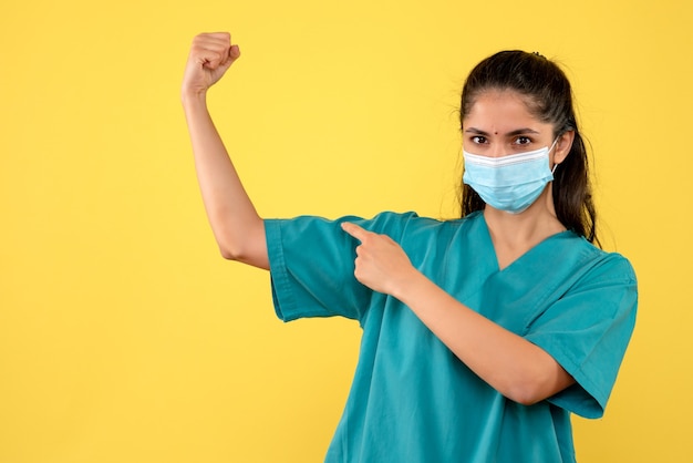 Front view of pretty female doctor with medical mask pointing at her arm muscle on yellow wall