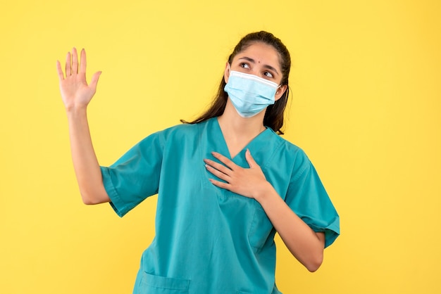 Front view of pretty female doctor with medical mask promising on yellow wall