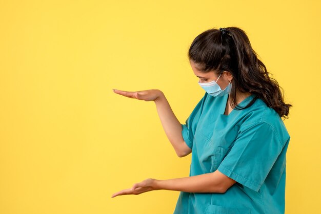 Front view of pretty female doctor with medical mask showing size with hands on yellow wall