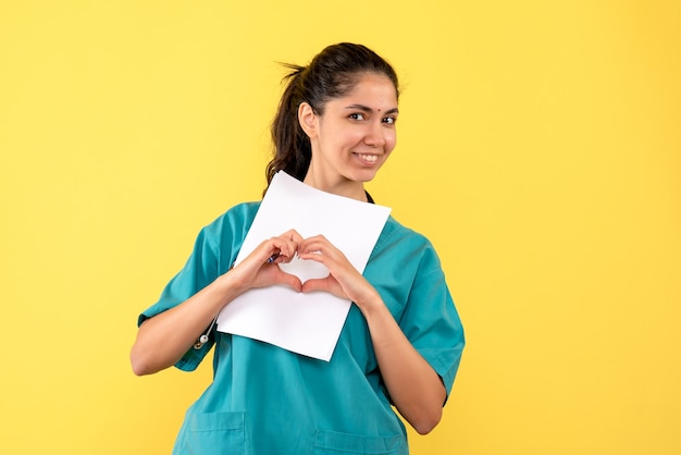 Free photo front view of pretty female doctor with papers making heart sign with hands on yellow wall