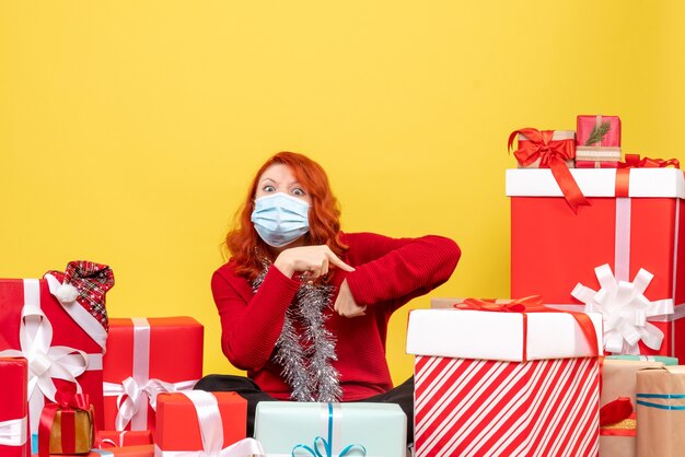 Front view pretty female sitting around presents in mask on yellow desk virus xmas new year covid- color