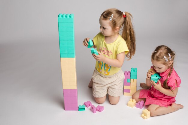 Front view of pretty sisters keeping hands together looking at camera and smiling on isolated background Lovely girls playing with building toys and enjoying game Concept of happiness and joy