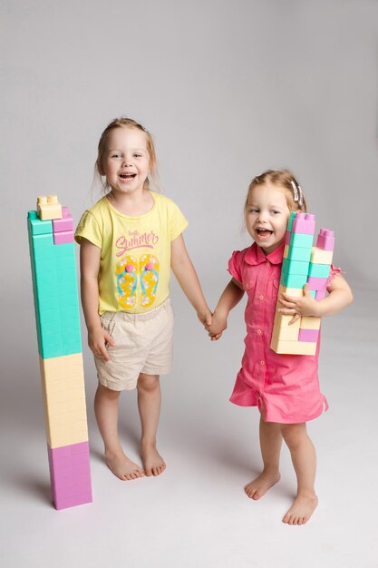 Front view of pretty sisters keeping hands together looking at camera and smiling on isolated background Lovely girls playing with building toys and enjoying game Concept of happiness and joy